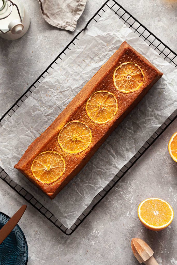 Orange cake with slice of orange on top, milk jar and pieces of orange lying on the sides.