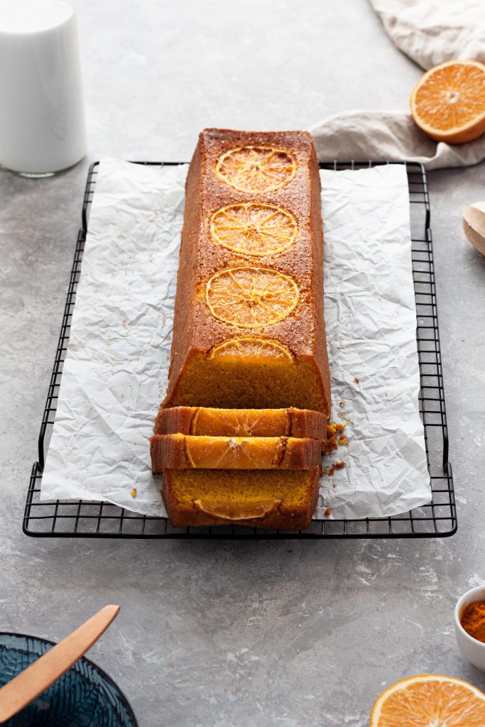 A loaf of orange upside-down cake with slices falling off, orange half lying in the background.