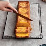 Hand slicing a loaf of orange tumeric cake with milk jar and halved orange in the background.