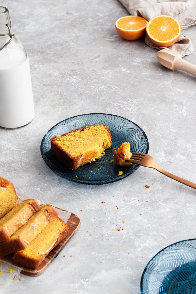 Slice of orange cake lying on a plate with milk jar on the side and rest of the cake in the background.