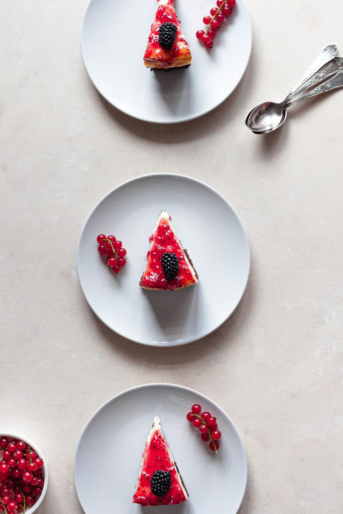 Three slices of white chocolate cheesecake served on plates, decorated with fruit syrup and fresh fruit.