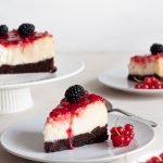 A slice of white chocolate cheesecake on a plate with raspberry sauce, decorated with fresh fruit.