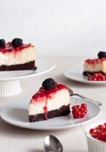 A slice of white chocolate cheesecake on a plate with raspberry sauce, decorated with fresh fruit.