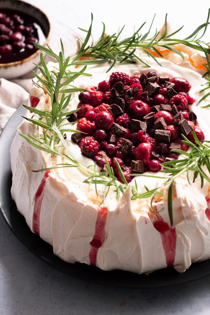 Decorated pavlova cake, topped with fresh rosemary, mascarpone cream, stone fruit and chocolate chunks.