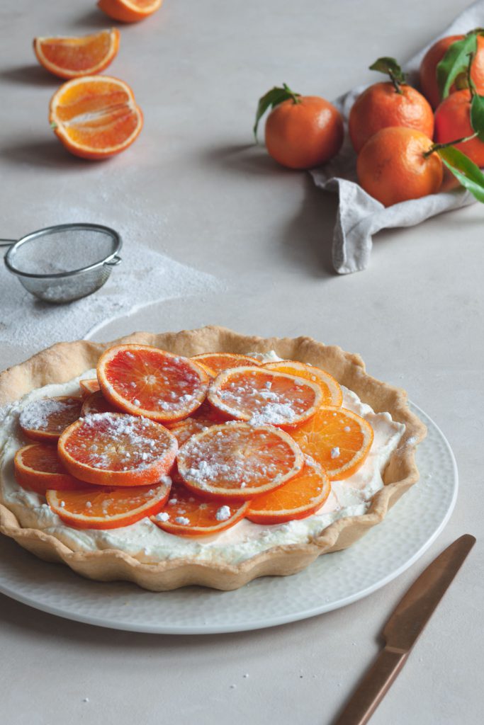 Tart decorated with fresh fruit and icing sugar, mandarines with leaves in the background.